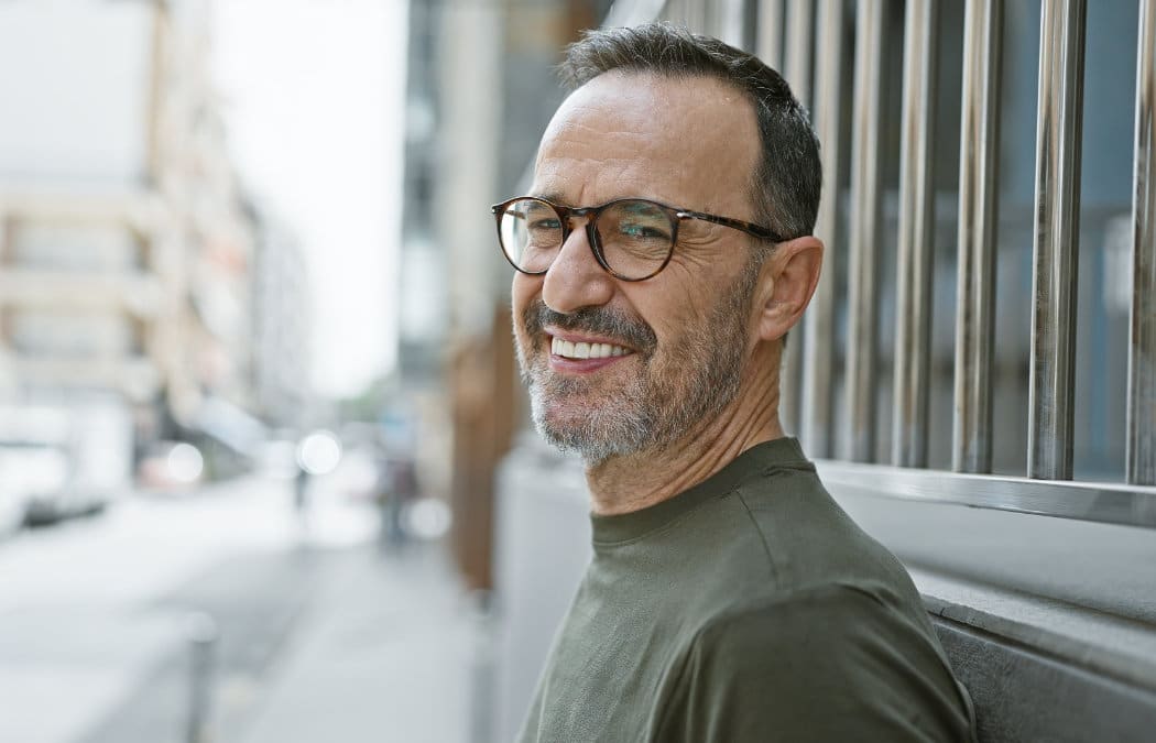 Middle-aged man with glasses and a beard standing outside by a metal fence, smiling at the camera on a city street.