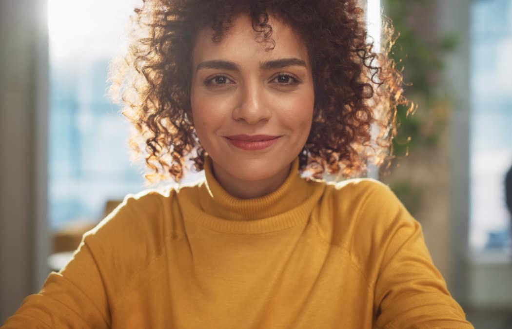 A person with curly hair and a yellow turtleneck sweater smiles while sitting indoors with sunlight in the background.