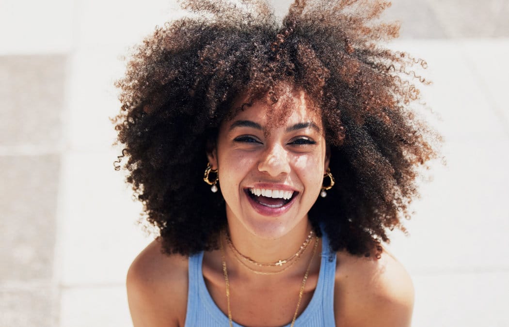 A person with curly hair, wearing a blue tank top and layered necklaces, smiles outdoors in bright sunlight.