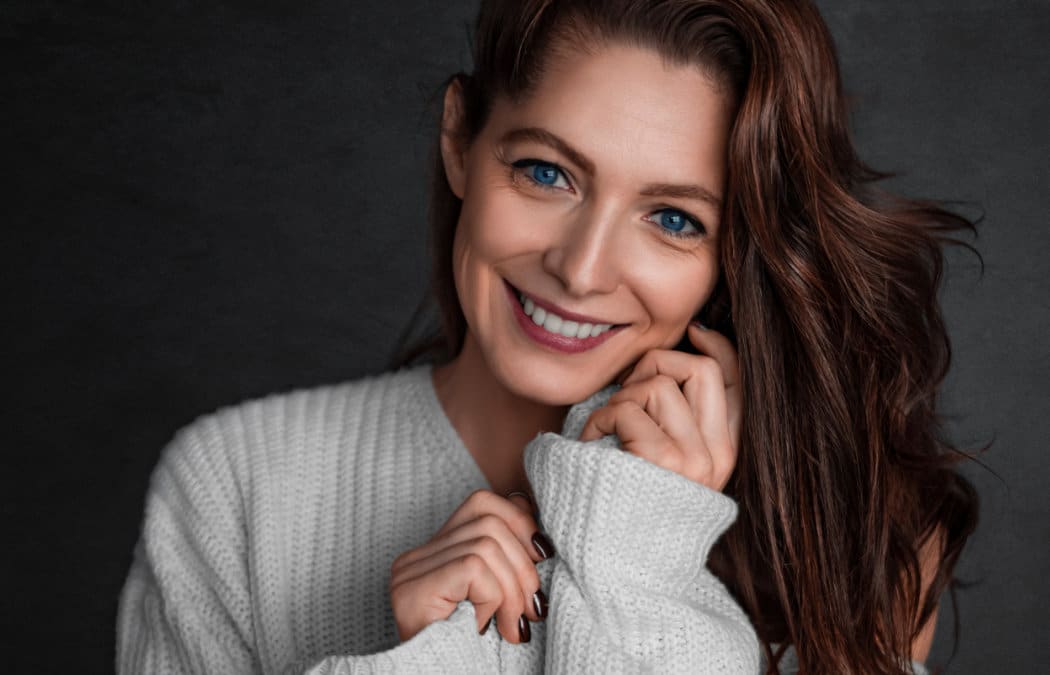 Woman with long brown hair and blue eyes smiles while wearing a white knit sweater against a dark background. She holds the sweater close to her face with both hands.