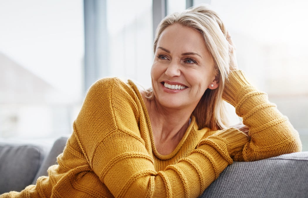 A woman in a yellow sweater sits on a couch, smiling and looking to her left with her hand resting on her head.